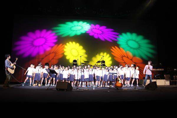 子供達が歌っている後ろでは、赤紫・黄色・オレンジ・緑と綺麗なお花の映像が映し出されている写真